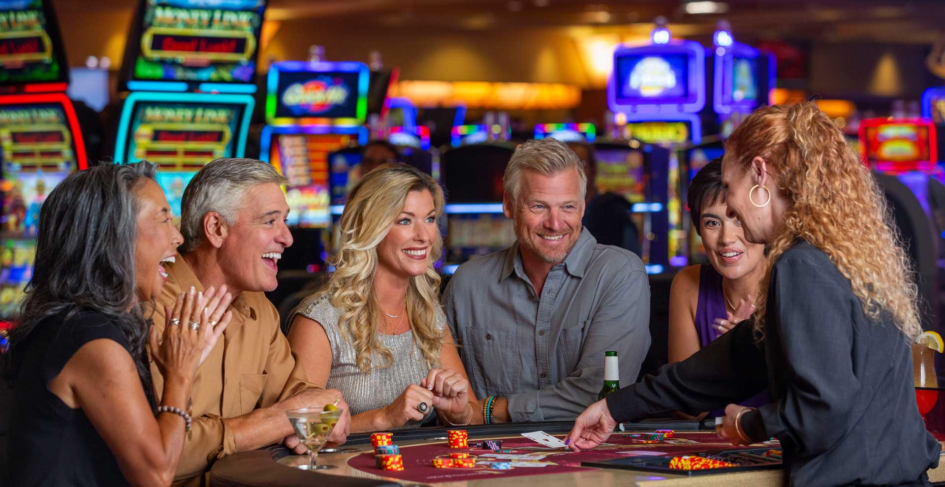 A group of five adults sitting around a casino table, interacting with a female dealer. The table has dice and poker chips, with colorful slot machines visible in the background. The individuals appear engaged and are smiling at one another.